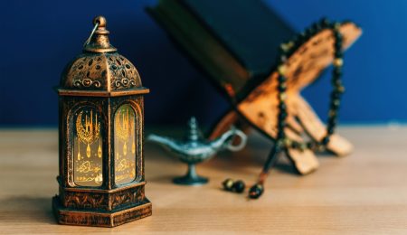 Lanterne ramadanesque traditionnelle avec calligraphie islamique, Coran ouvert sur un repose-livre en bois, chapelet musulman et lampe décorative sur fond bleu – Préparatifs du Ramadan 2025 en Tunisie.