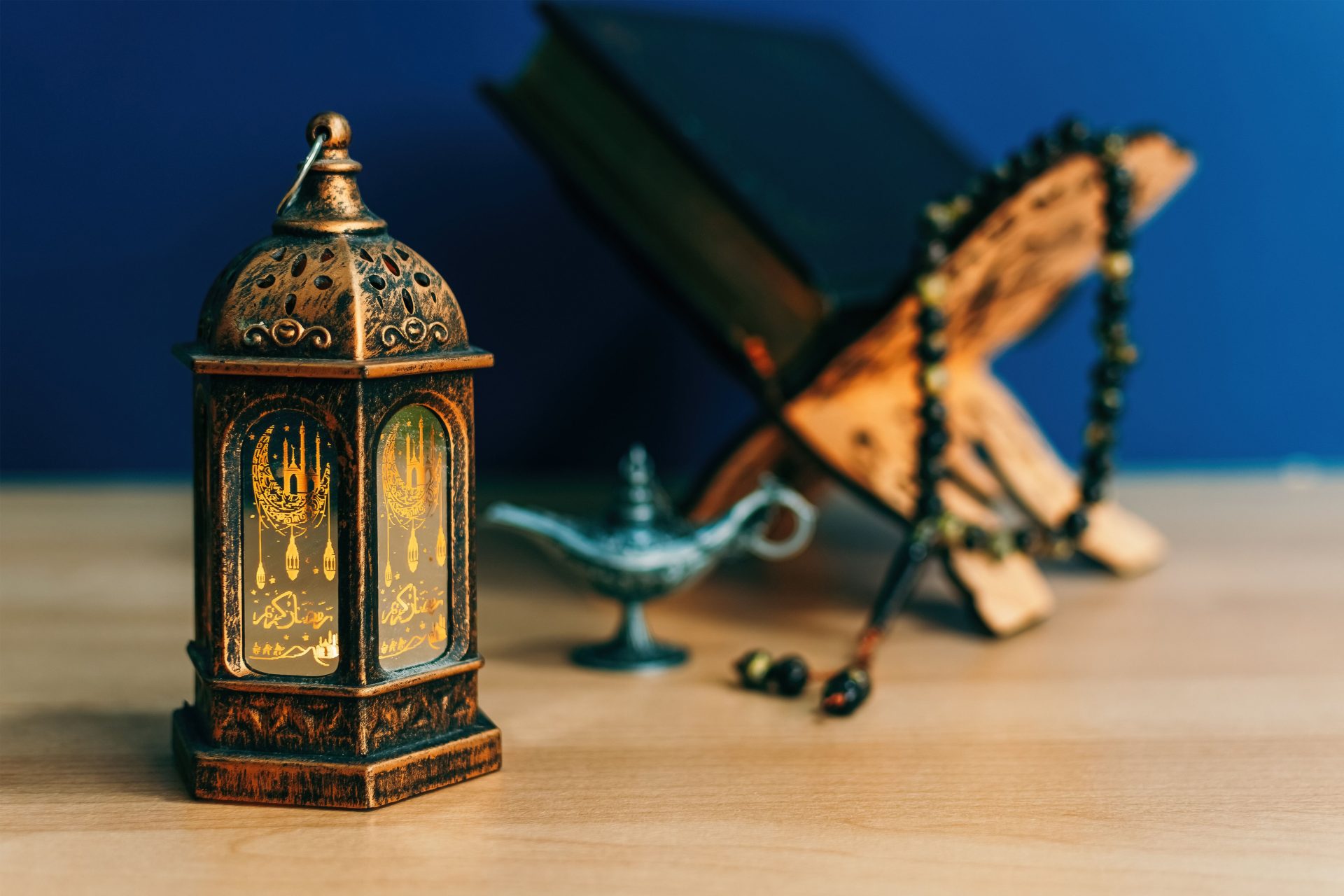 Lanterne ramadanesque traditionnelle avec calligraphie islamique, Coran ouvert sur un repose-livre en bois, chapelet musulman et lampe décorative sur fond bleu – Préparatifs du Ramadan 2025 en Tunisie.