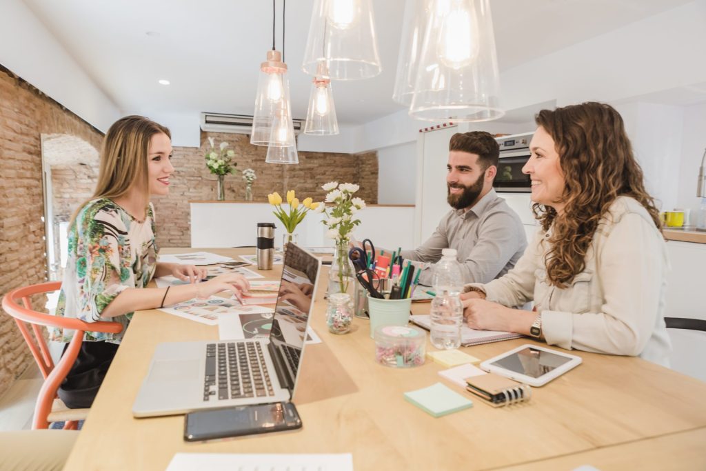 Trois collègues souriants collaborent autour d'une table avec ordinateurs portables, documents et matériel de bureau dans une cuisine moderne