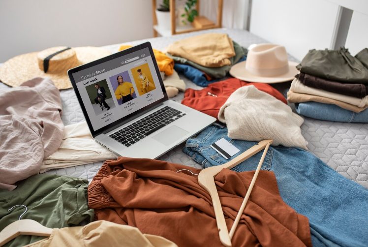Femme faisant du shopping en ligne sur ordinateur portable entourée de vêtements