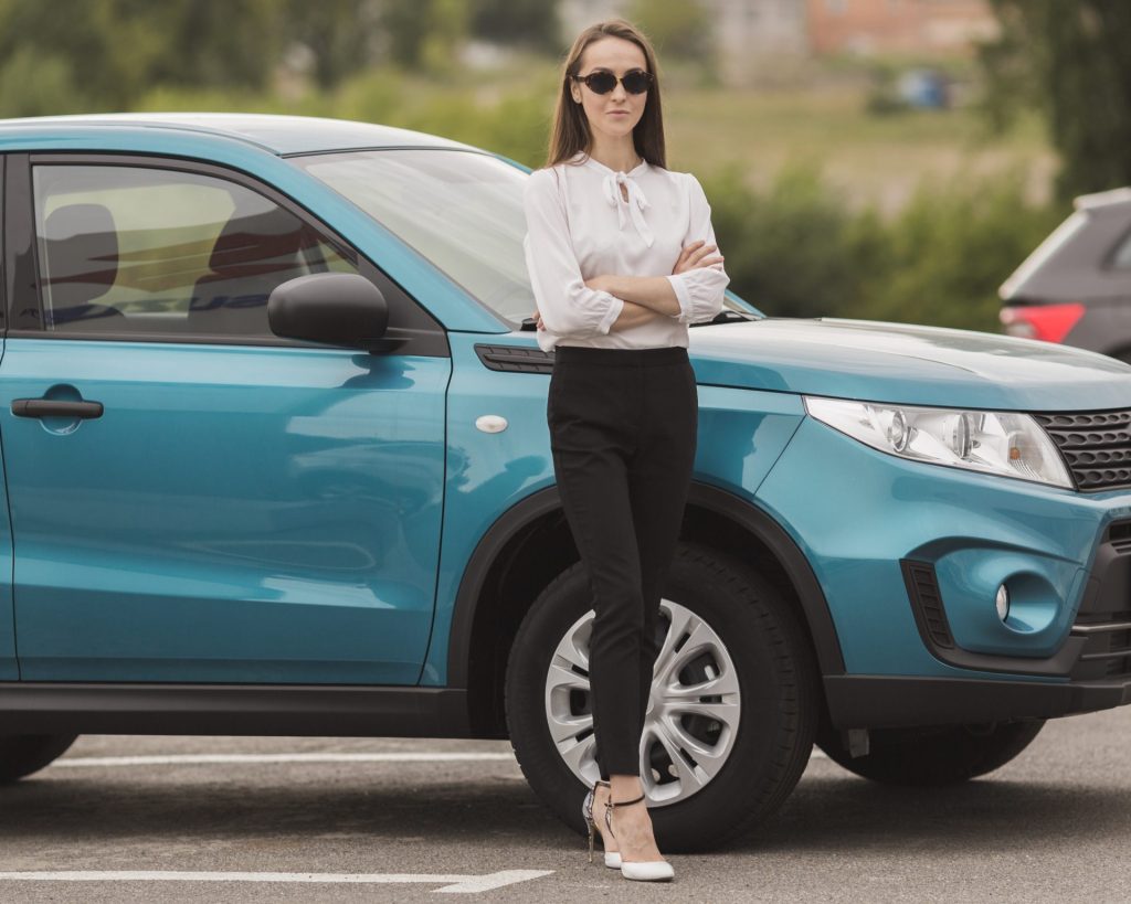 Femme en tenue professionnelle appuyée contre une citadine compacte bleue dans un parking